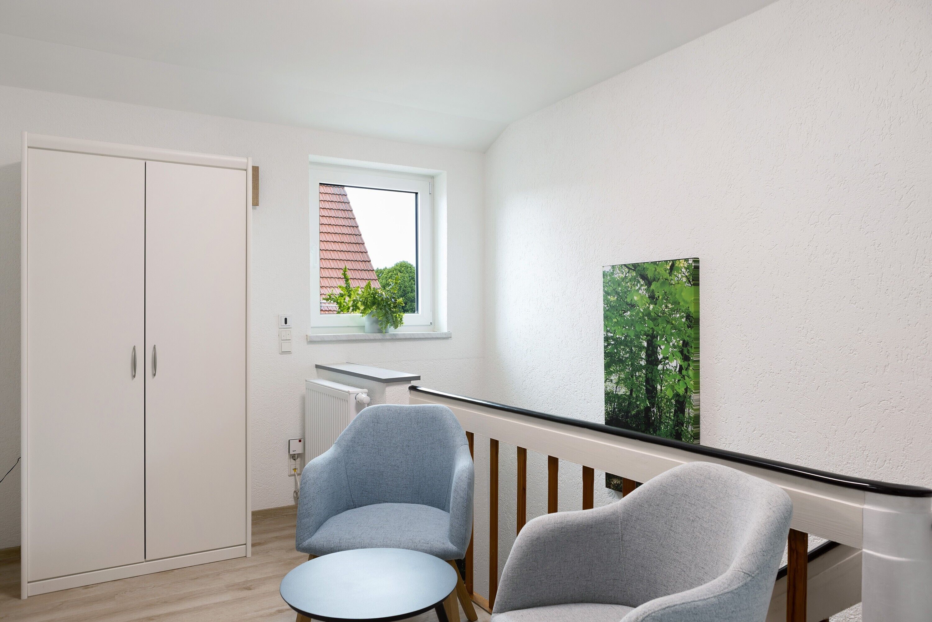 Een kamer met een witte garderobe, een klein tafeltje, twee stoelen, een raam met uitzicht op bomen en een schilderij aan de muur.