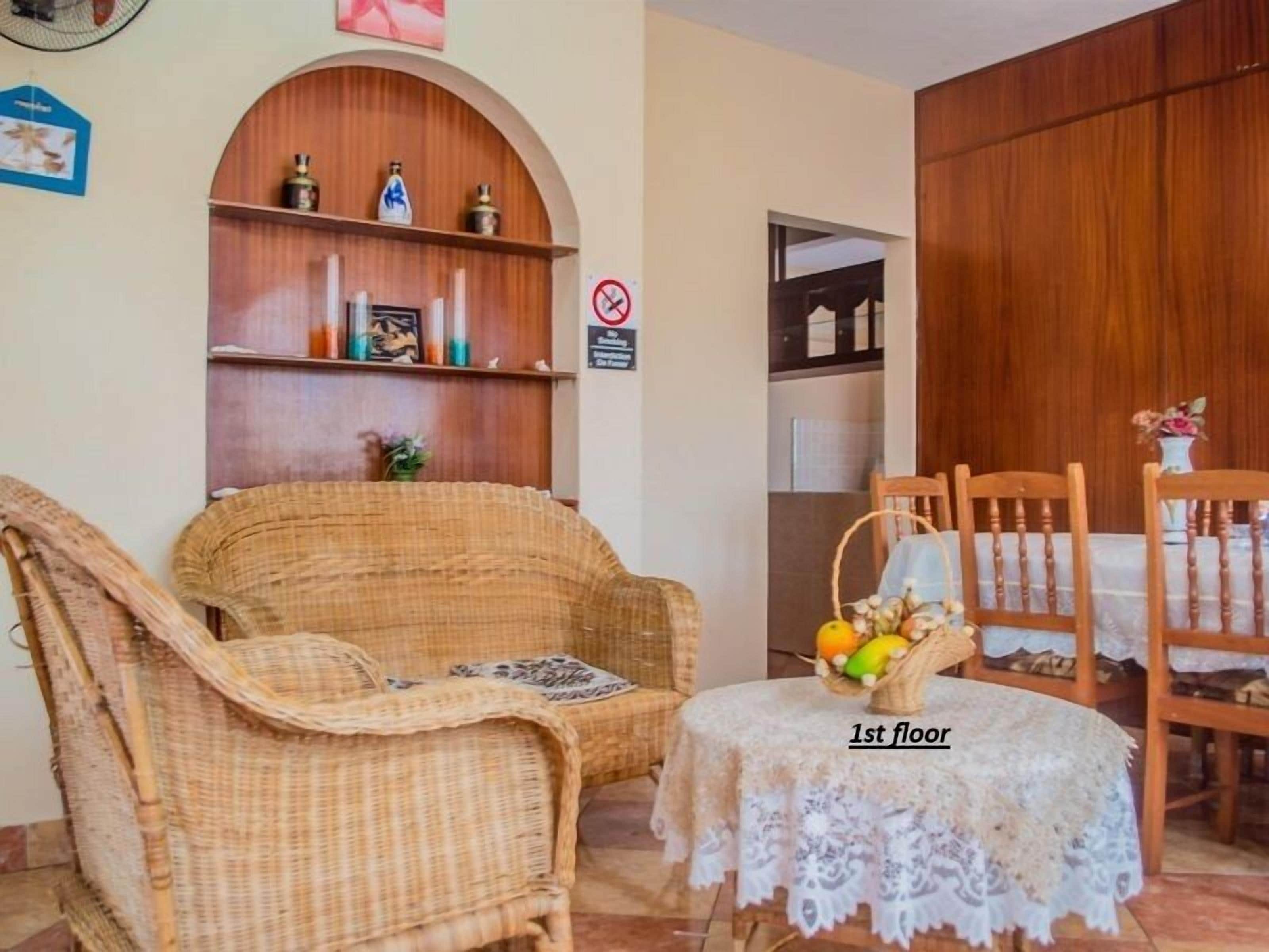 A room with a wicker sofa, a small table with a lace tablecloth, and a dining area with wooden chairs and a basket of fruit.