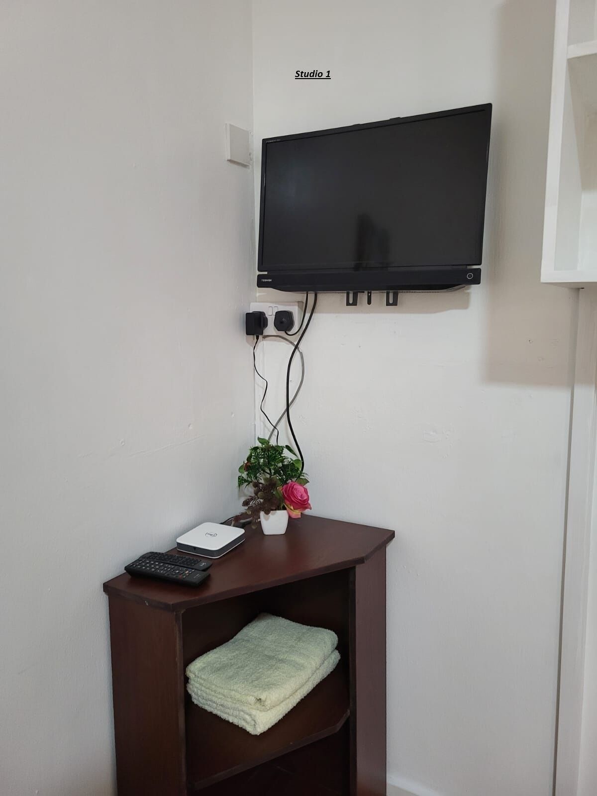 A room with a mounted TV, a desk with a laptop, a keyboard, a plant, and a folded towel.