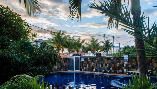 A swimming pool surrounded by lush greenery, with a stone wall and a small structure nearby. - Villa Nass (Flic-en-Flac)