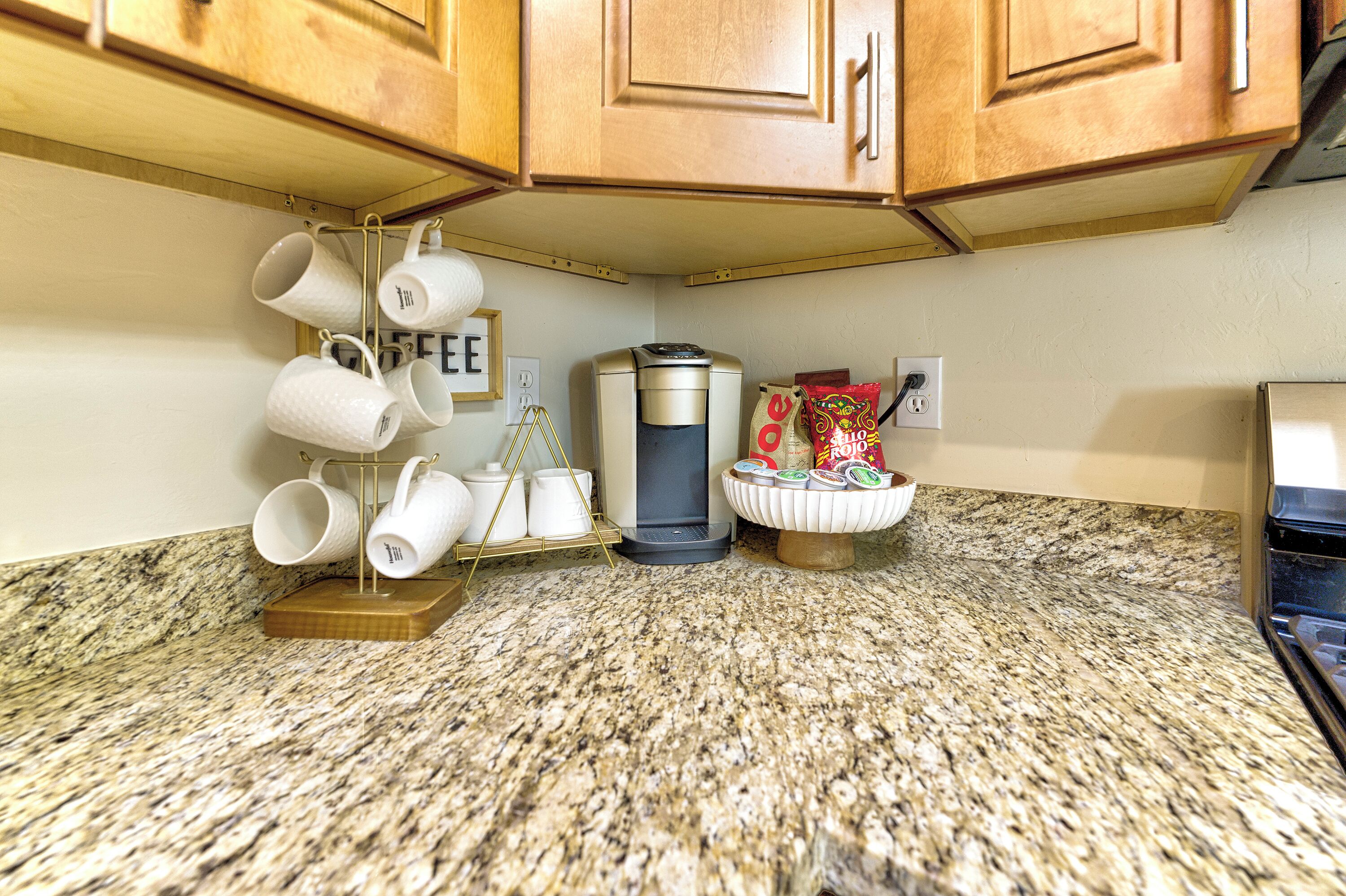 A kitchen with granite countertops, wooden cabinets, a coffee maker, and a mug rack.
