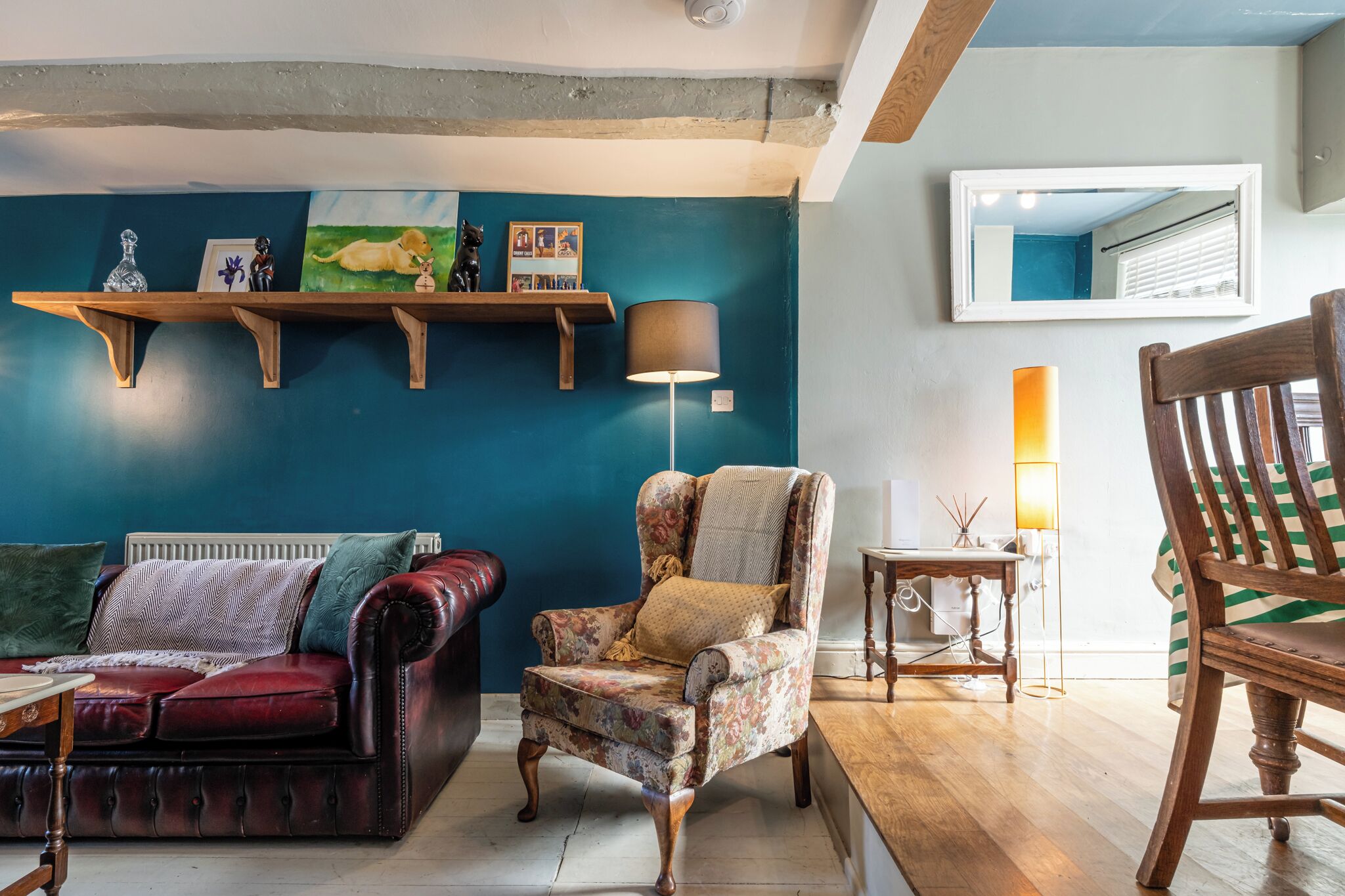 A living room with a dark blue wall, a red leather sofa, a floral armchair, and a wooden chair. There are shelves with decorative items and a floor lamp.