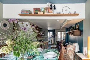 Un comedor con una mesa dispuesta para cuatro personas, una estantería arriba y una cocina al fondo.