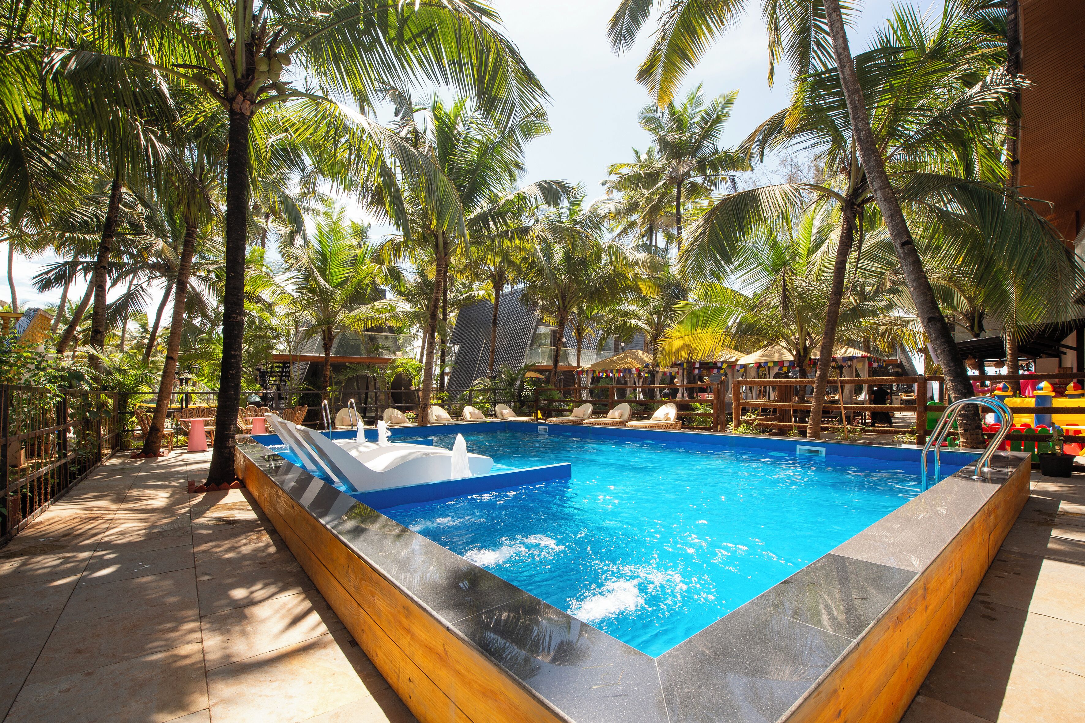 Une piscine rectangulaire dotée d’une terrasse en bois et agrémentée d’un élément décoratif aquatique bleu, le tout entouré de palmiers et de chaises longues.