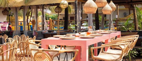 An outdoor dining area with wicker chairs and a pink table, surrounded by thatched umbrellas and hanging light fixtures.