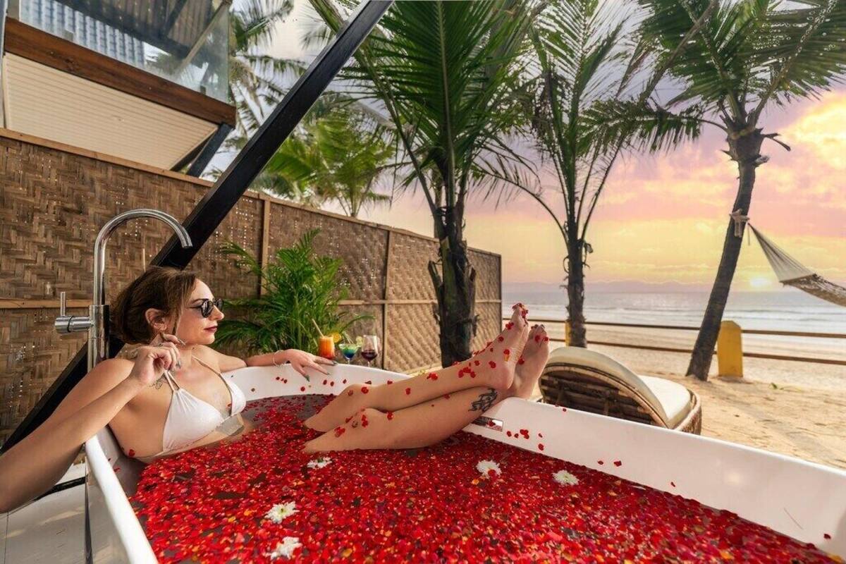 Una mujer se relaja en una bañera llena de pétalos de rosa, con una playa y palmeras al fondo.