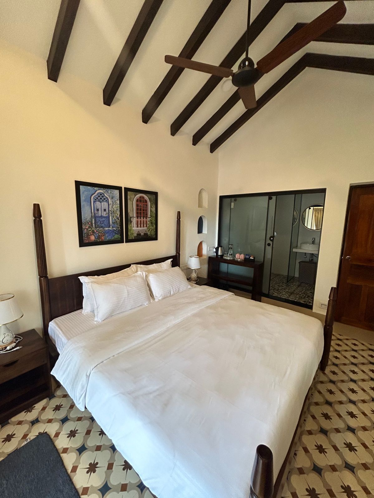A bedroom with a large bed, a ceiling fan, two framed pictures on the wall, a wardrobe, and a bathroom visible through a glass door.