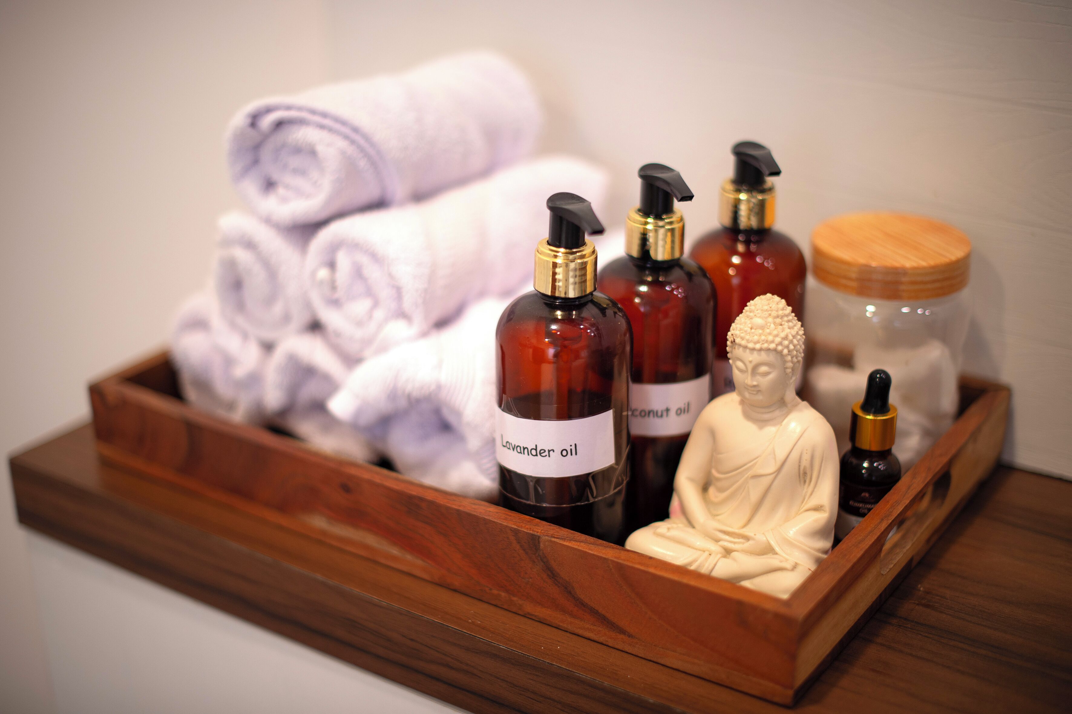 Una bandeja de madera con toallas enrolladas, botellas de aceite de lavanda, aceite de coco y una estatua de Buda.