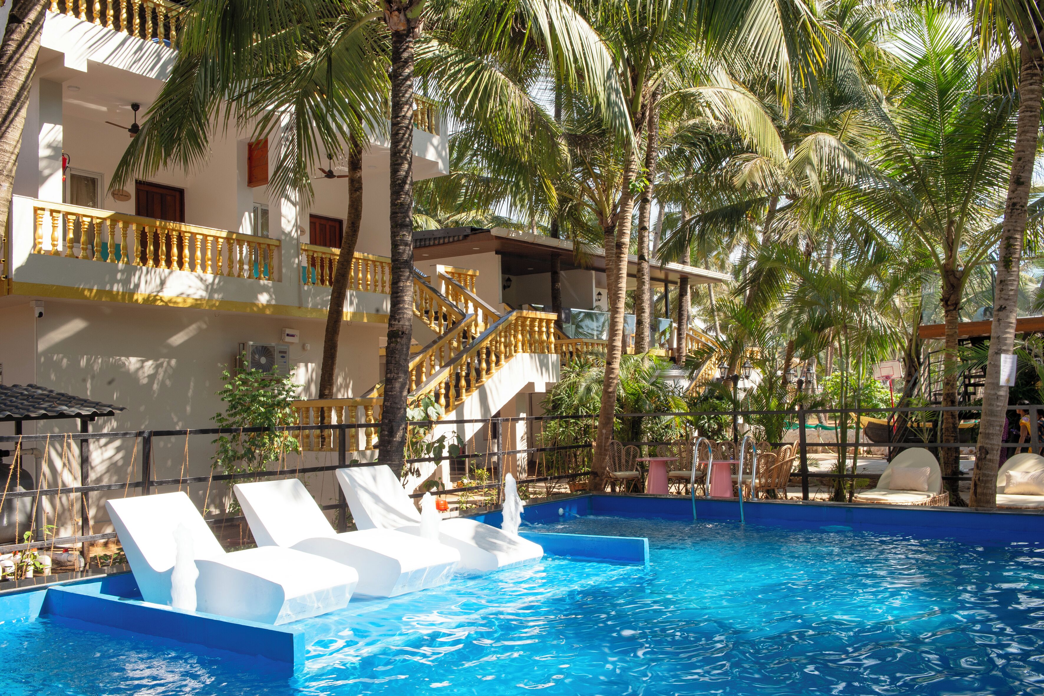 A swimming pool with white lounge chairs, palm trees, and a multi-story building with balconies.