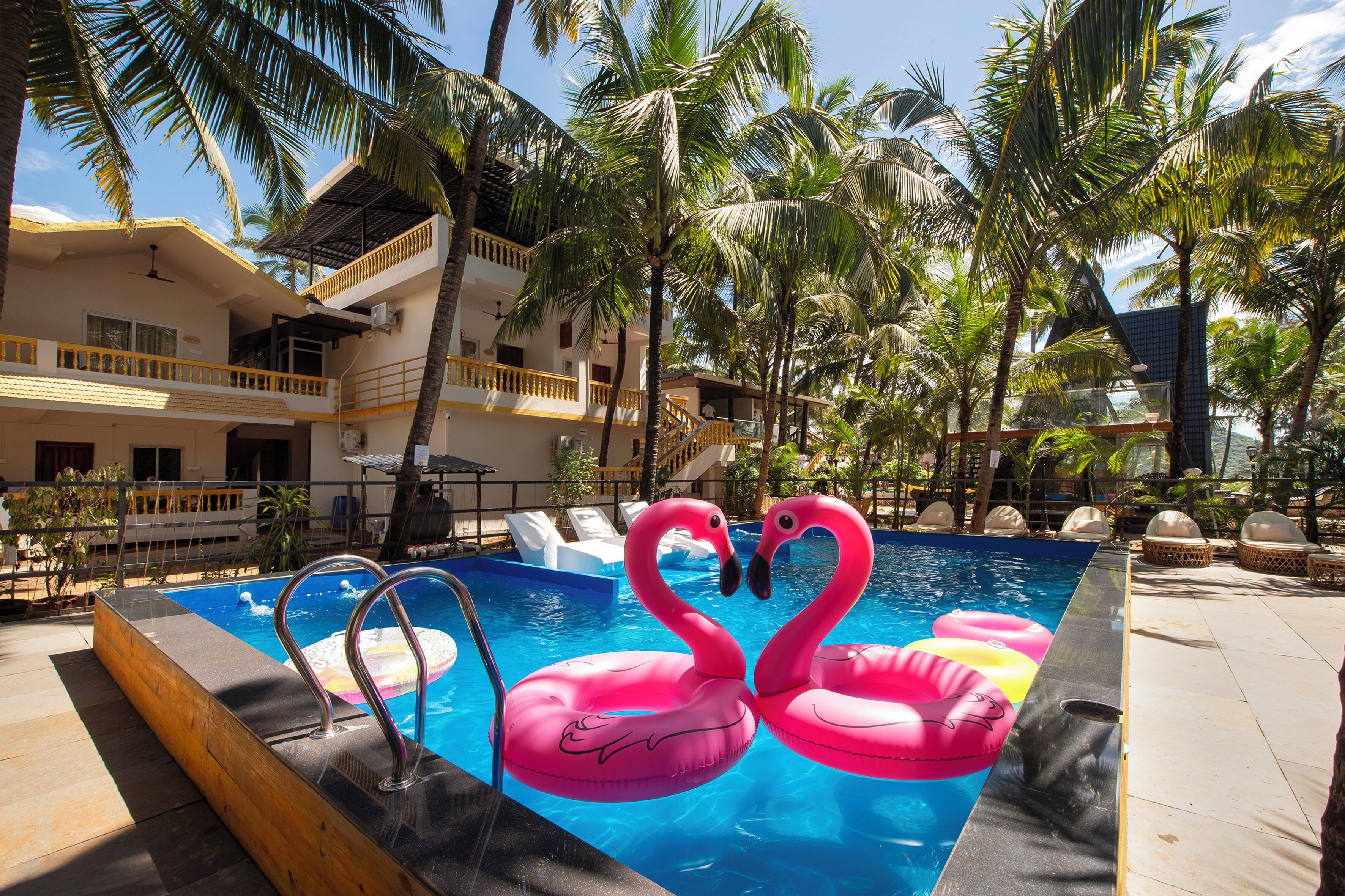 Una piscina con flotadores con forma de flamenco rosa, rodeada de palmeras y un edificio de varios pisos con balcones.