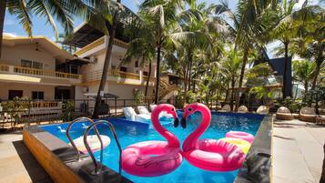 Una piscina con flotadores con forma de flamenco rosa, rodeada de palmeras y un edificio de varios pisos con balcones.
