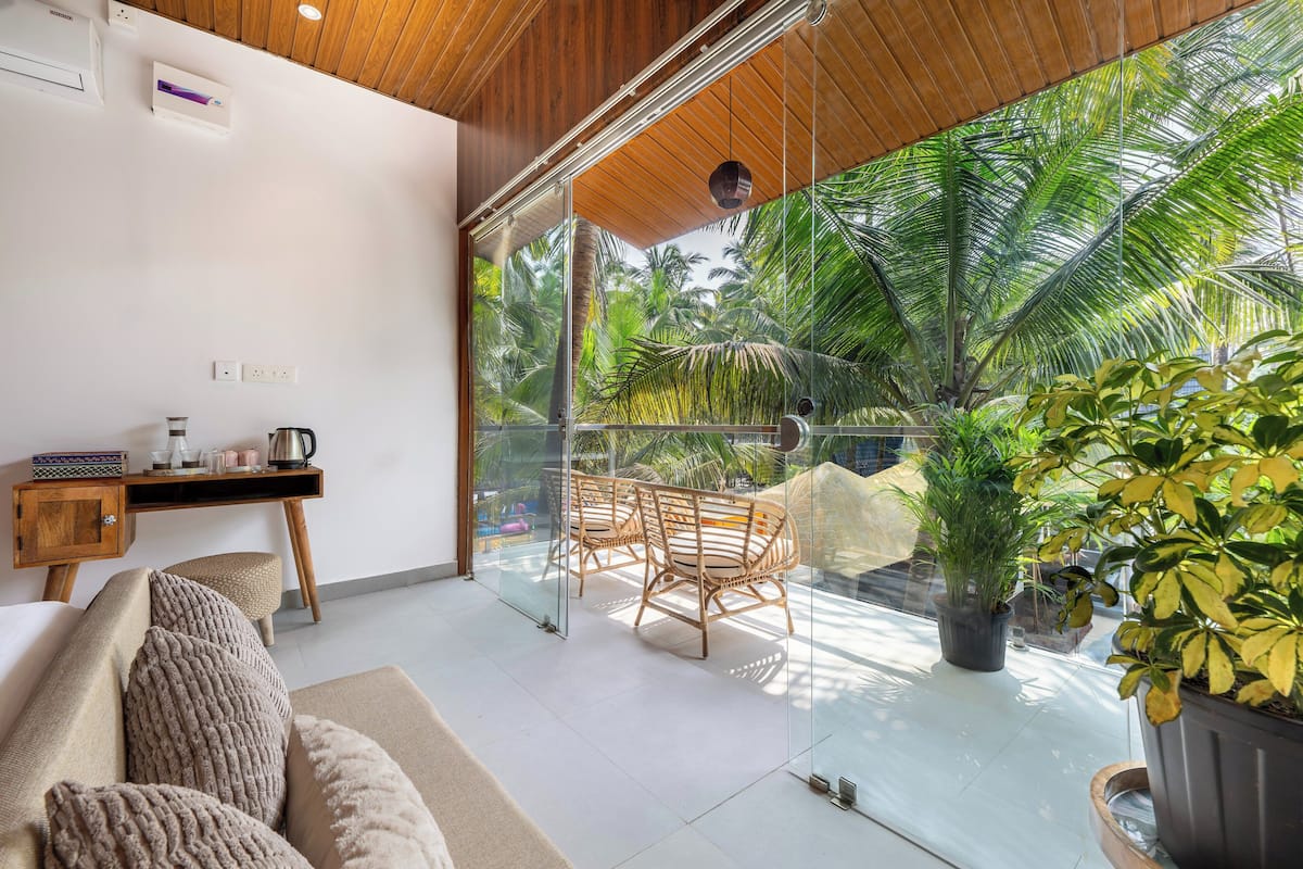 Una habitación moderna con pared de vidrio, techo de madera, un sofá con cojines texturizados, una pequeña mesa de madera con un hervidor y vistas a palmeras.
