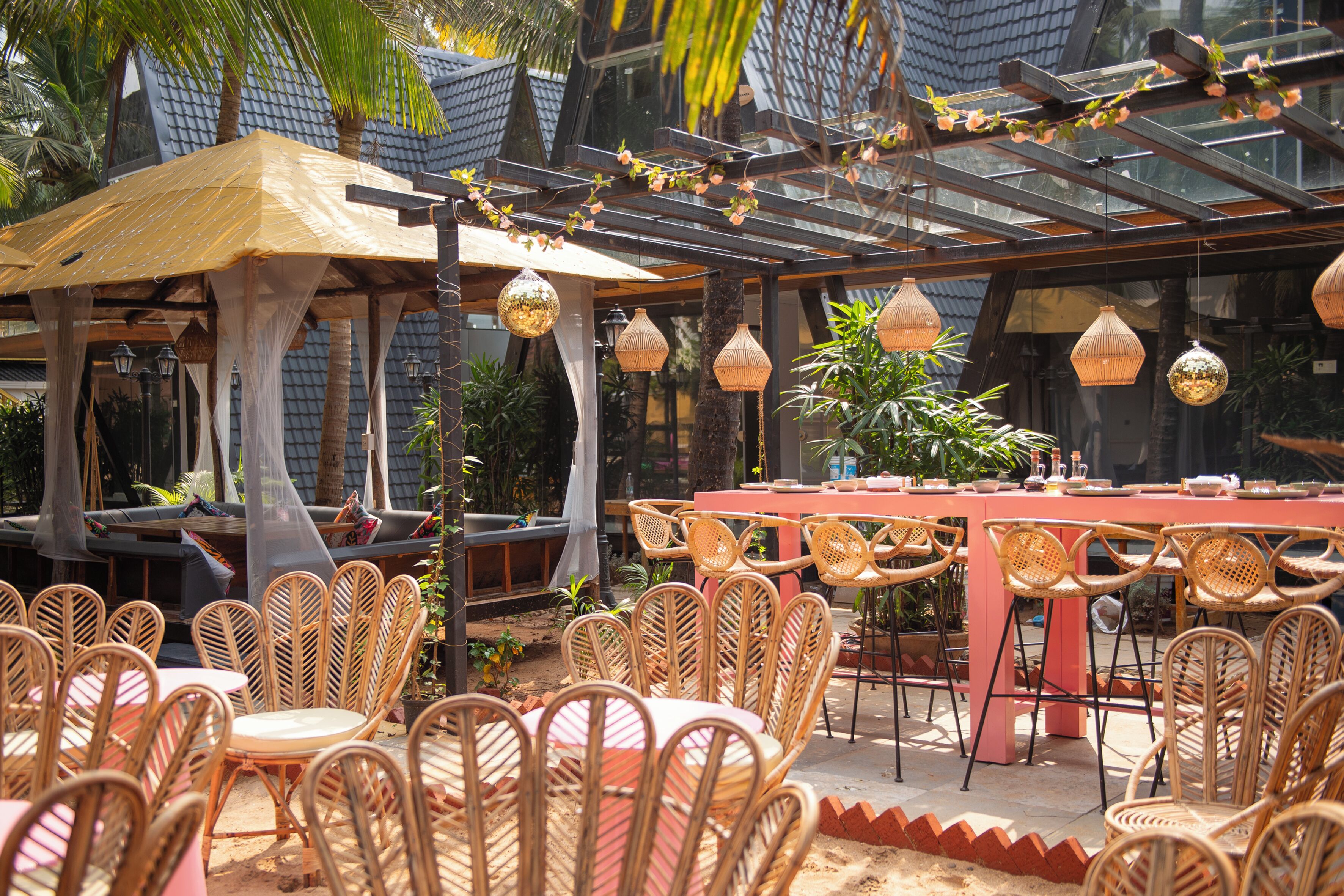 Un espace de restauration en plein air avec des chaises et des tables en osier, une pergola illuminée de guirlandes lumineuses et un plafond en verre.