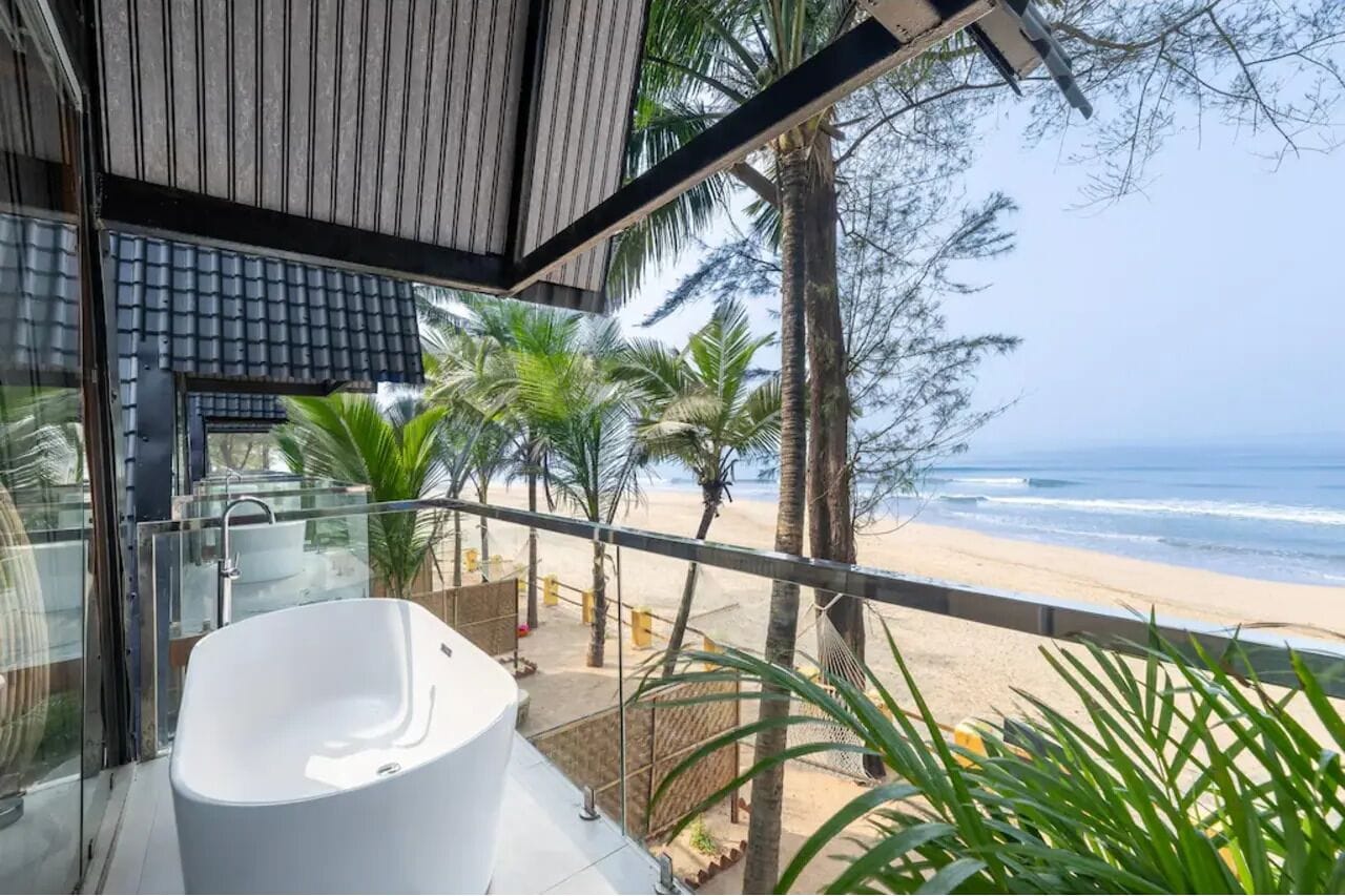 Varanda moderna com uma banheira branca, com vista para uma praia com palmeiras e o oceano.