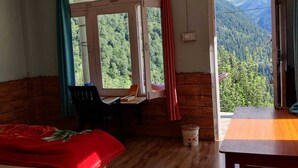Habitación con cama, escritorio y mesa. Hay dos ventanas con cortinas, una con vista a una cadena montañosa cubierta de bosque.