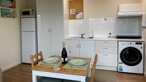Een praktische keuken met een magnetron, koelkast, spoelbak en wasmachine. Een eettafel met stoelen en een fles champagne.