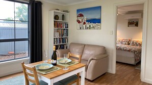 Een gezellige woonkamer met een beige bank, een houten eettafel gedekt voor twee personen, een boekenplank vol boeken, en een schilderij van een kustlandschap.