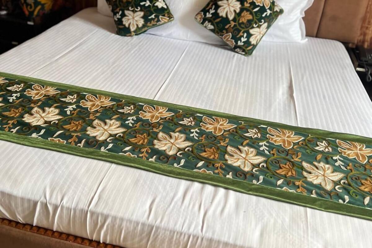 A bed with a white sheet, two decorative pillows with a floral pattern, and a green embroidered runner.