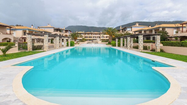A swimming pool with a stone border, surrounded by buildings with a Mediterranean architectural style, set against a backdrop of hills and a cloudy sky. - Villa Blu Mare Relax Place in Nafpaktos! (NAFPAKTOS)