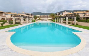 Una piscina con borde de piedra, rodeada de edificios de estilo arquitectónico mediterráneo, frente a colinas y un cielo nublado.