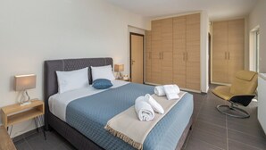 A neatly made bed with a blue bedspread and a beige throw blanket, a wooden wardrobe, a chair, and a bedside table with a lamp. - Villa Blu Mare Relax Place in Nafpaktos! (NAFPAKTOS)