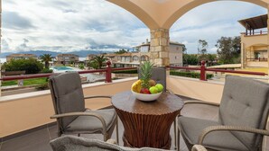 A balcony with wicker chairs, a round table with a fruit bowl, and a view of a pool and buildings. - Villa Blu Mare Relax Place in Nafpaktos! (NAFPAKTOS)