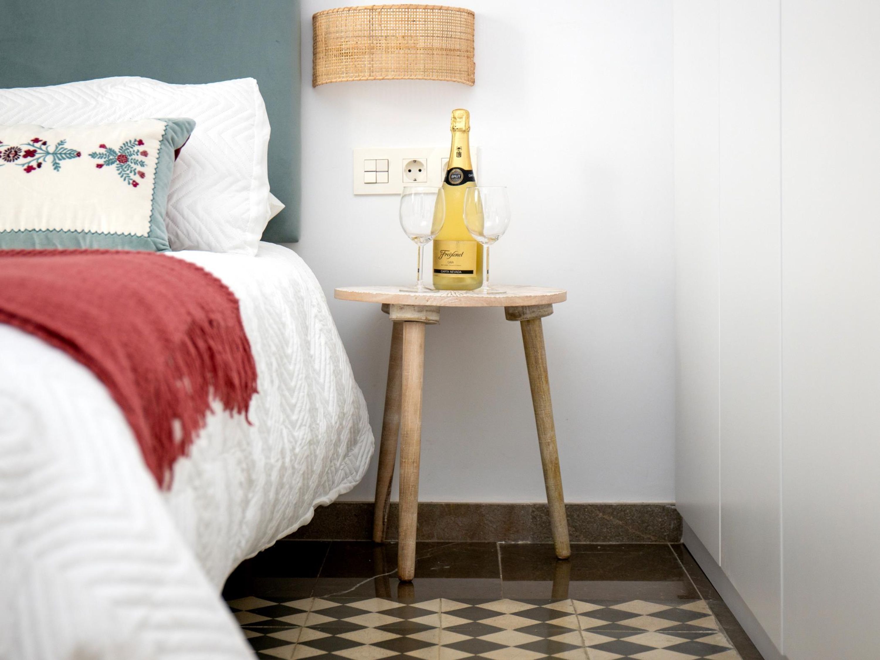 Una cama bien tendida con una manta blanca y roja, una pequeña mesa de madera con una botella de champán y una copa, y un enchufe de pared.