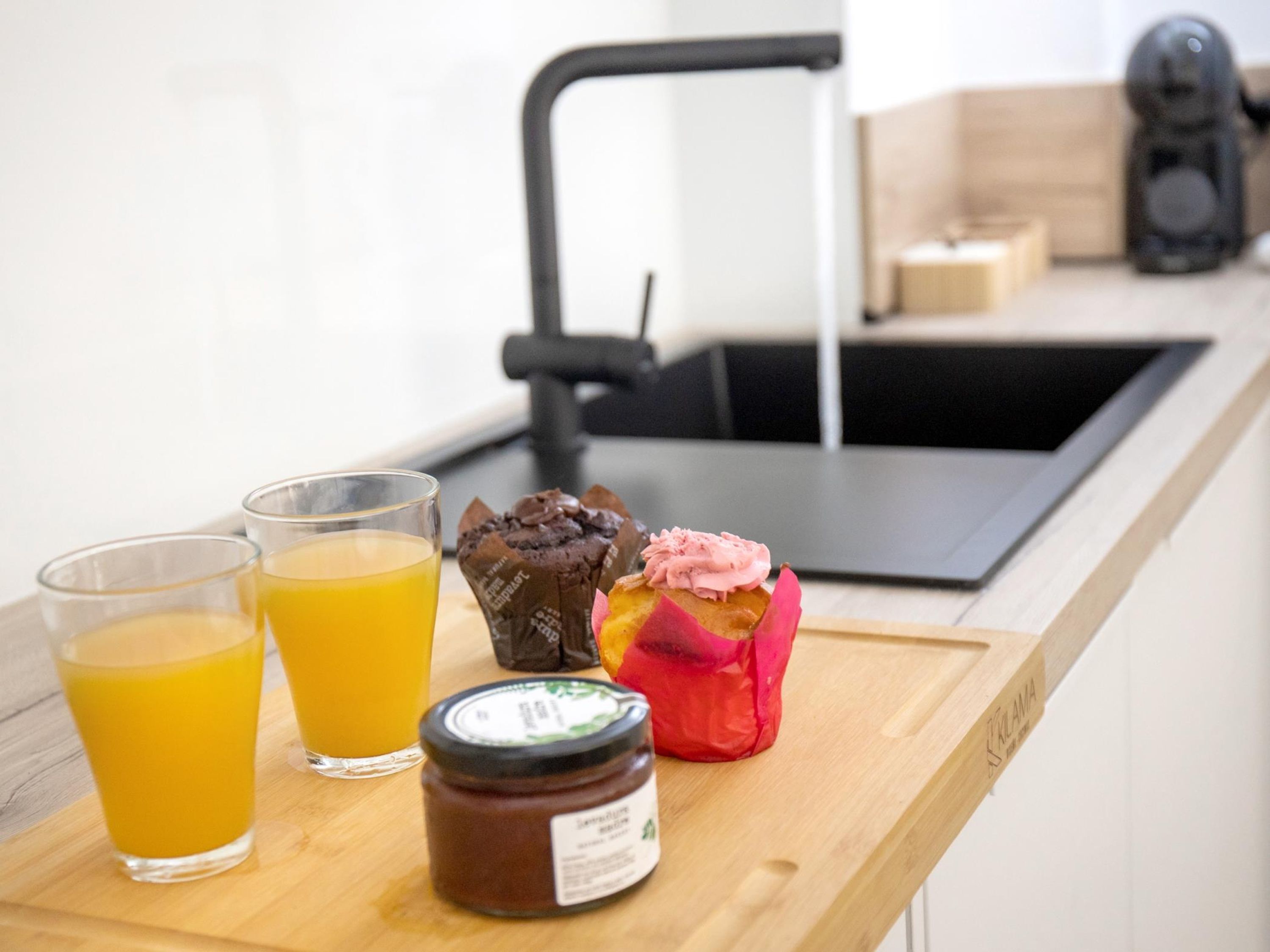 Ein Frühstücksbrett mit zwei Gläsern Orangensaft, einem Muffin mit rosa Zuckerguss und einem Marmeladenglas auf einer Holzauskidde in einer Küche.
