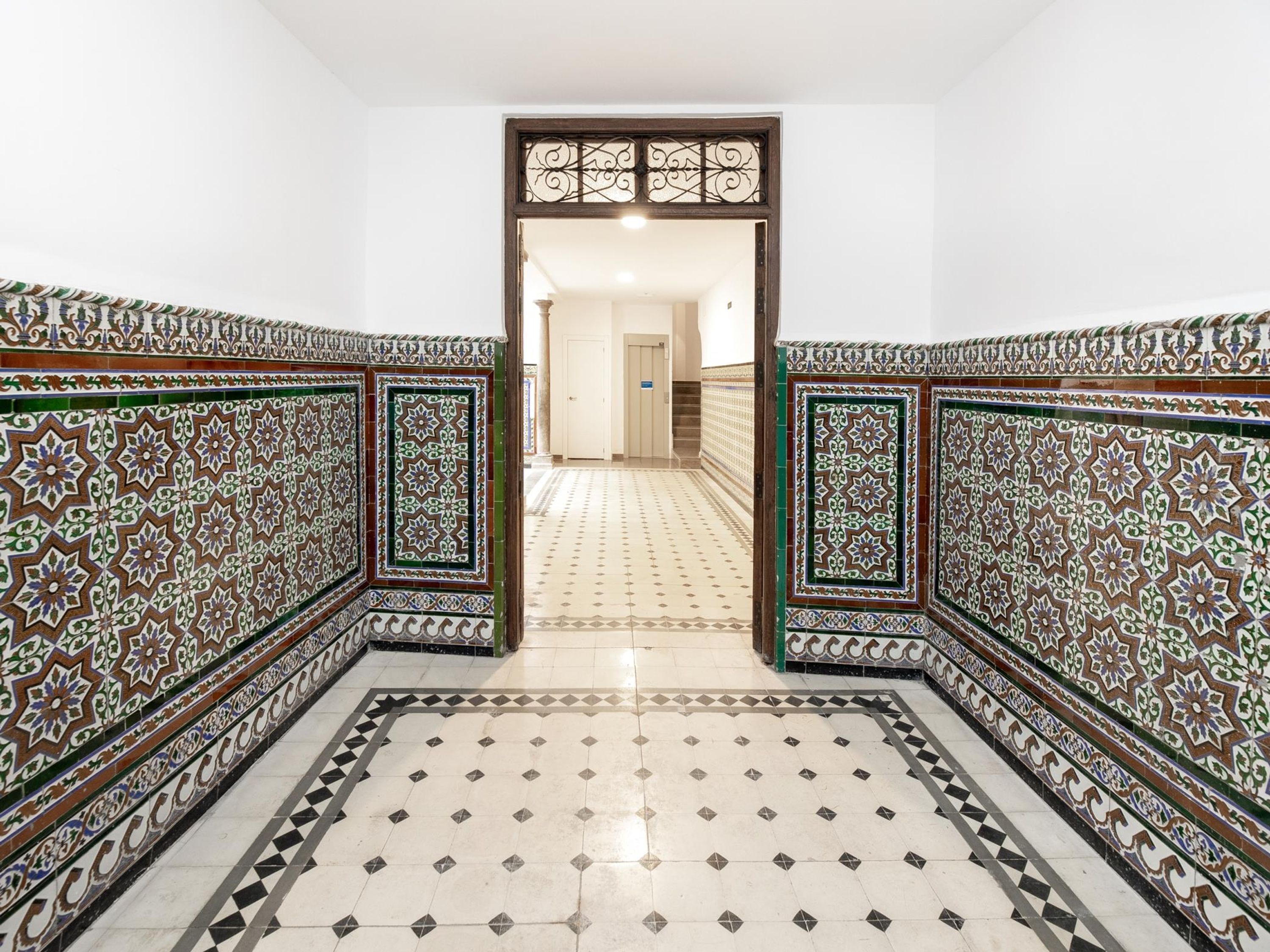 A hallway with intricate tile patterns on the walls and floor, leading to a door at the end.