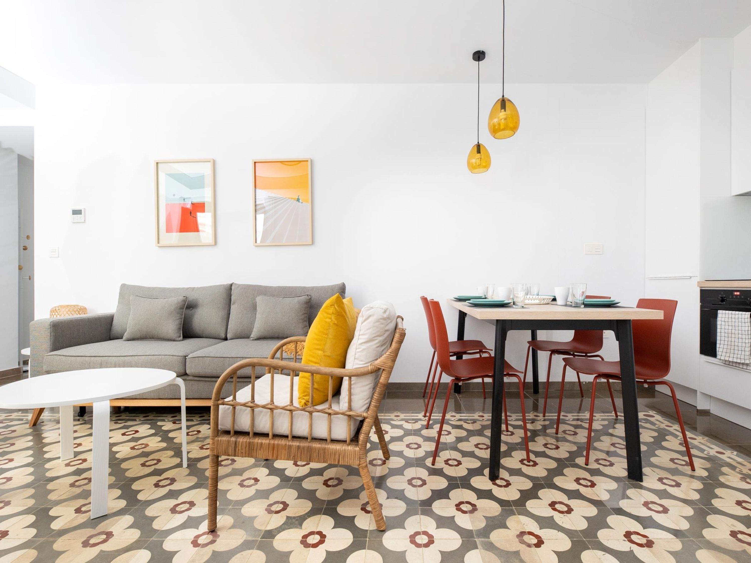 A modern living room with a grey sofa, a wicker chair, a dining table with red chairs, and a patterned floor.