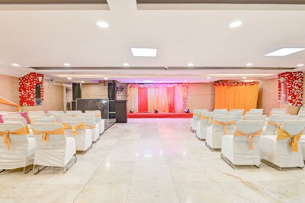 A conference room with rows of white chairs with orange sashes, a stage with a red curtain, and a podium. - Hotel Marks INN & Banquet (Noida)