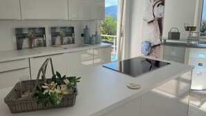 A modern kitchen with white cabinetry, a central island with a built-in induction cooktop, and a large window offering a view of greenery and mountains. - Modern vacation apartment with view and pool near Baden Baden in the Black Forest  (Gernsbach)