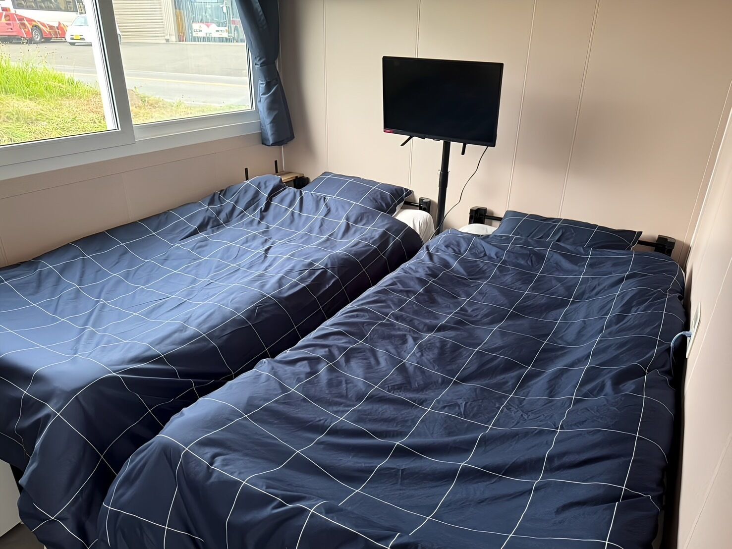 Two single beds with blue bedding, a TV on a stand, and a window with a view of the street.