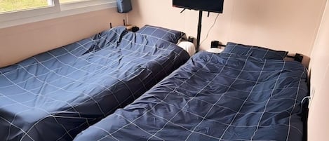 Two single beds with blue bedding, a flat-screen TV mounted on the wall, and a window with curtains.
