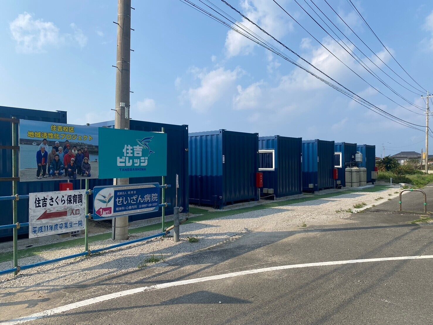 A row of blue portable toilets with a sign for Tanegashima in the background.
