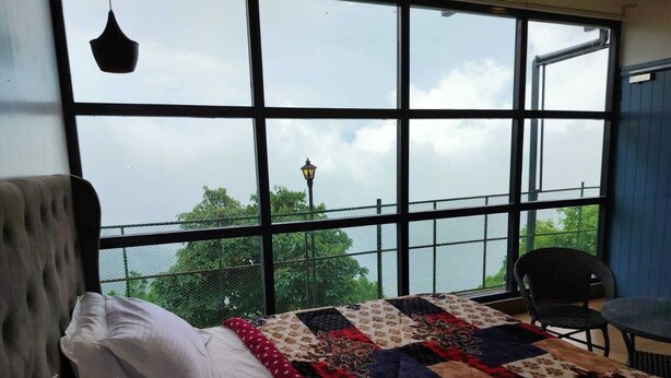 Een slaapkamer met een groot raam, een bed met wit beddengoed, een kleurrijke quilt en een stoel bij het raam.