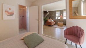 A bedroom with a bed, a green pillow, a pink chair, a shower, a staircase, and a living area with a couch and a plant. - Holiday home with Jacuzzi 200m from the beach. (Wemeldinge)