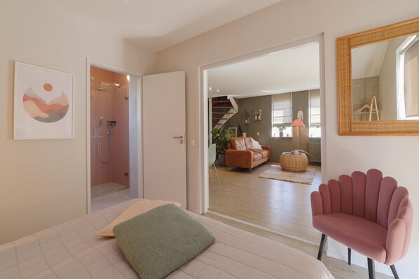 A bedroom with a bed, a green pillow, a pink chair, a shower, a staircase, and a living area with a couch and a plant.