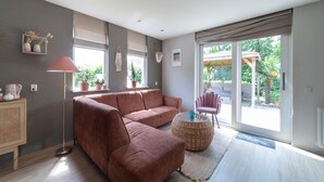 A modern living room with a sectional sofa, a floor lamp, and a round coffee table. There are potted plants on the windowsill and a view of the outdoors through large sliding glass doors. - Holiday home with Jacuzzi 200m from the beach. (Wemeldinge)