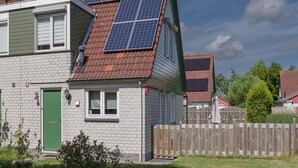 Una casa de dos pisos con techo inclinado, paneles solares, una puerta verde y una valla de madera.