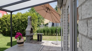 Un patio techado con una parrilla de piedra, una mesa de concreto y una planta en maceta con flores rosadas.