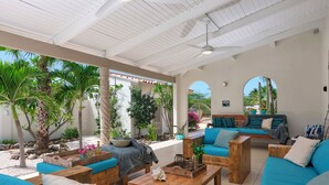 Patio esterno con mobili in legno, una ventola a soffitto e vista su un giardino con cactus e palme.