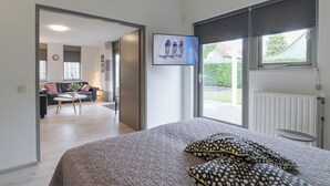 A bedroom with a bed, a TV mounted on the wall, a window with a view of the outside, and a door leading to a living area with a sofa and a coffee table. - Holiday home: Bubbles, Bounce and Relax (Wemeldinge)