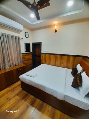 A bedroom with a bed, wooden headboard, ceiling fan, and a clock on the wall. - Amma Rooms and Dormitory (Raipur)