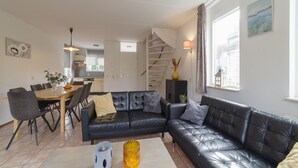 A modern living room with a black leather sofa, a dining table with chairs, and a staircase leading to an upper level. - Holiday Home in Zeeland with Garden (Wemeldinge)