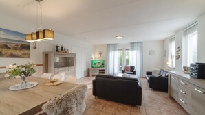 A modern living room with a sofa, a dining table with a vase of flowers, a television, and a kitchen area with cabinets and a countertop.