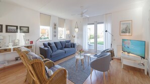Un salon moderne avec un canapé bleu, des fauteuils en osier, une table basse, un téléviseur et de grandes fenêtres.