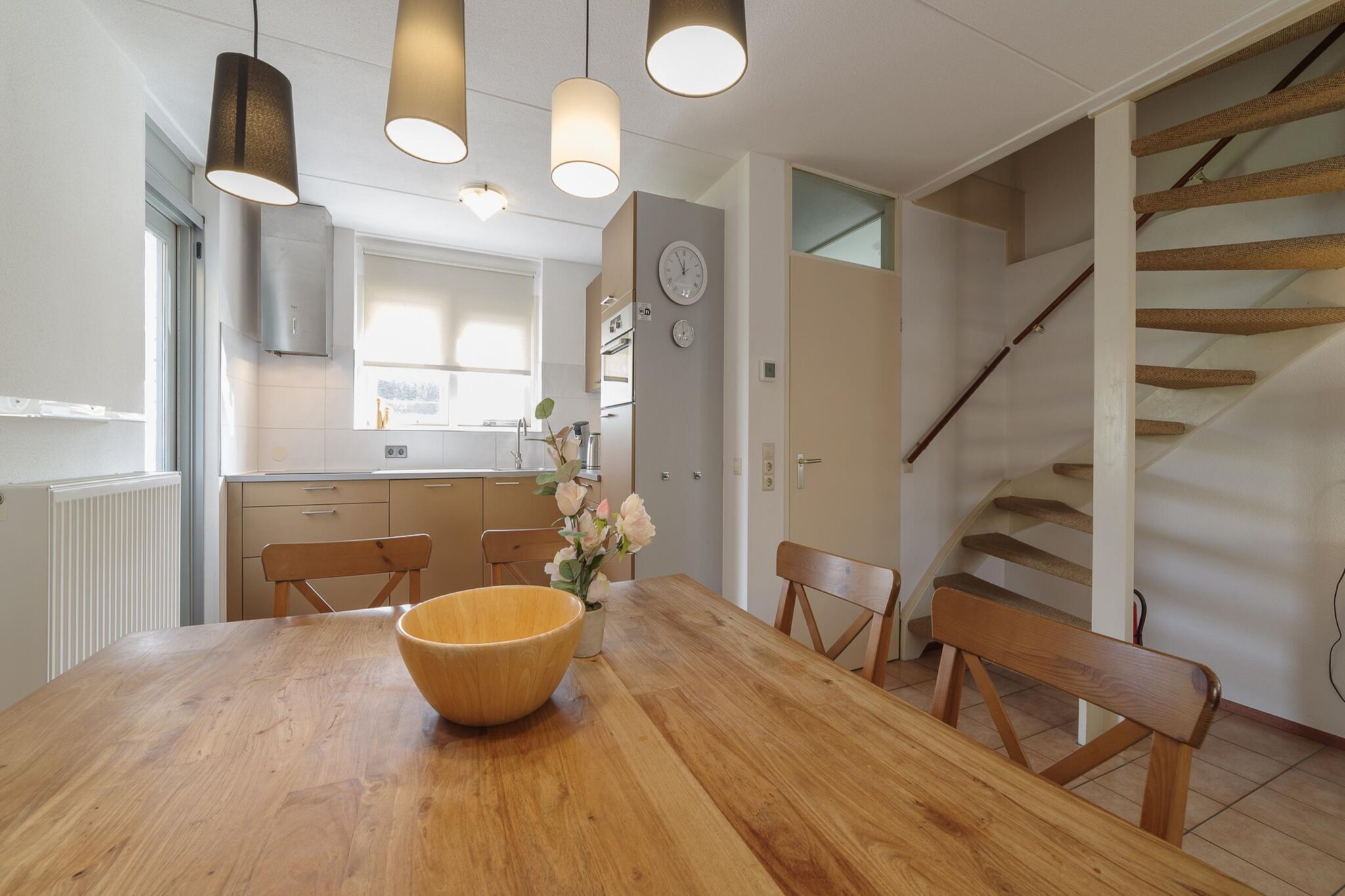 A modern kitchen with a wooden dining table, hanging pendant lights, and a clock on the wall.