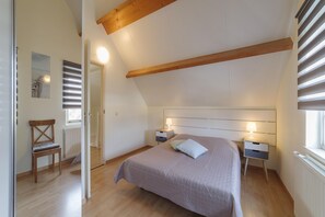 Un dormitorio con techo inclinado, vigas de madera, una cama con cabecera, dos mesitas de noche con lámparas, una silla y un radiador.