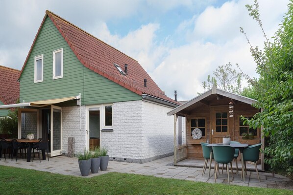 Een huis met twee verdiepingen, met een groen dak en witte muren, en een terras met tuinmeubilair en potplanten.